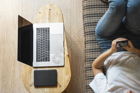 女人躺在沙发上使用智能手机 桌上放着笔记本电脑和电子阅读器 在线学习 自由职业 个体经营者或自由职业者的女孩使用电话 并在笔记本背景图片