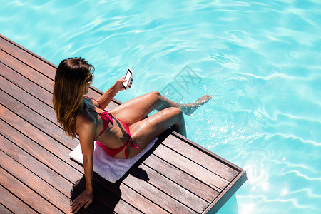 沙滩装在游泳池边缘使用智能手机的妇女闲暇享受游泳衣假期家庭生活电话水池晴天技术泳装背景