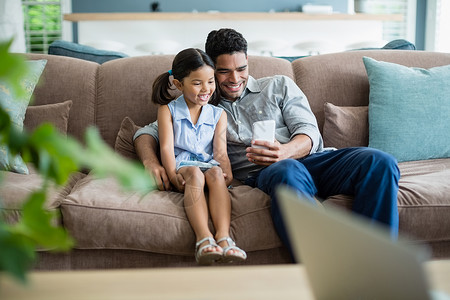 一位父亲和女儿父亲和女儿坐在沙发上 使用手机家庭生活家庭童年服装孩子住所女性沟通女孩男性背景