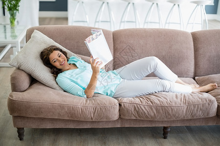 微笑的女人在客厅沙发上躺在沙发上时看杂志图片