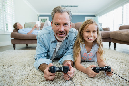 一位父亲和女儿快乐的父亲和女儿躺在起居室地板上玩电玩游戏时休闲微笑房子乐趣休息娱乐服装游戏现实喜悦背景