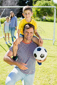 跪舔父亲带着足球微笑 他的父亲在公园上肩并肩背着儿子去打橄榄球背景