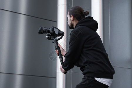 年轻的专业摄像师在 3 轴万向稳定器上手持专业相机 专业设备有助于制作高质量视频而不会抖动 身穿白色连帽衫的摄影师正在制作视频博背景