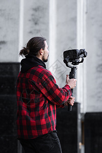 年轻的专业摄像师在 3 轴万向稳定器上手持专业相机 专业设备有助于制作高质量视频而不会抖动 摄影师穿着红色衬衫制作视频背景