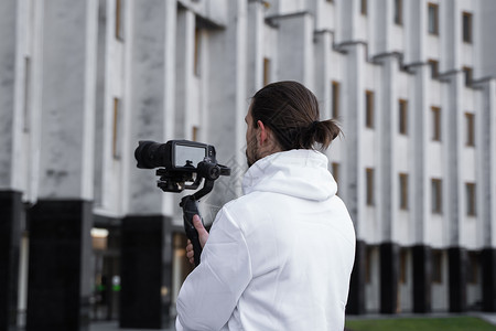 年轻的专业摄像师在 3 轴万向稳定器上手持专业相机 专业设备有助于制作高质量视频而不会抖动 身穿白色连帽衫的摄影师正在制作视频录背景