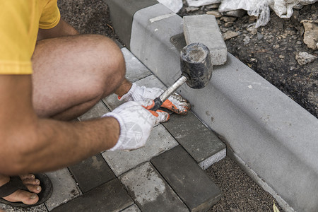 铺路砖一名建筑工人的手戴着橙色手套 手上拿着锤子配件 正在铺设新的外部铺路石 小心地将一块铺路石放在水平和倾斜的土壤基础上 沙地基 建背景