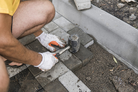 铺路砖一名建筑工人的手戴着橙色手套 手上拿着锤子配件 正在铺设新的外部铺路石 小心地将一块铺路石放在水平和倾斜的土壤基础上 沙地基 建背景