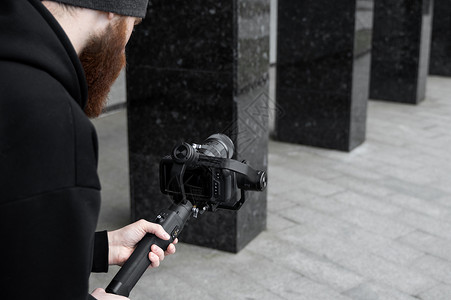 身穿黑色连帽衫 留着胡子的专业摄像师 手持专业相机 安装在 3 轴万向稳定器上 电影制作人使用专业的电影摄影机制作精彩视频 电影背景