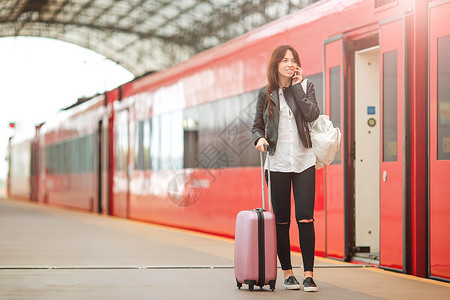 红色手提箱携带行李的年轻女子在火车站用手机交谈 她旅行时乘快车等候客服旅游者 笑声乘客铁路火车电话旅游游客车站平台车皮速度背景