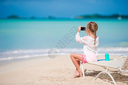 小女孩照片小女孩用照相机拍摄录像或照片 坐在日晒上海滩支撑假期手机微笑旅游幸福互联网热带游客背景