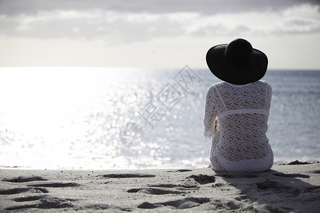 坐在海边留着长发的年轻女子在黎明时分在风中看着地平线 身着白色蕾丝连衣裙 白色内衣和大黑帽海景帽子女性天空戏剧性孤独悲伤身体生活背景图片