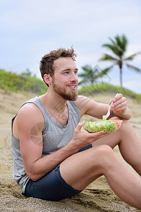 吃冰棒沙滩男子健身男子在锻炼时吃健康的沙拉餐 英俊的年轻肌肉发达的成年男性在午休时间跑步锻炼后坐在沙滩上 新鲜准备去素食生蔬菜食品背景