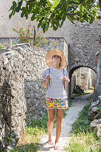 草帽图美丽的金发年轻女旅行者戴着草帽太阳帽享受地中海的夏天 在一棵无花果树下采摘水果 树上有薰衣草花 背景是传统的老石屋背景
