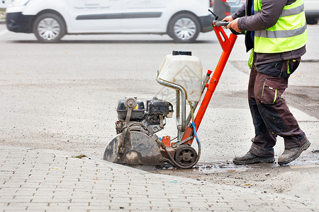 一名工人在城市街道的背景下 用车道上的煤气锯断了旧沥青行动人行道维修工作背心割机反光建设者淡绿色工具背景图片