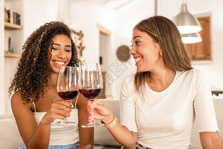 混合种族两位年轻的混血女性在家里用红酒杯庆祝 — 微笑着的西班牙裔卷发黑发女郎和她最好的朋友在她现代优雅的客厅里坐在他的沙发上叮当作响背景