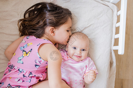 可爱的抱抱和亲吻她快乐的妹妹 在床上喜悦小姑娘家庭裙子闲暇蝴蝶黑发孩子内饰乐趣背景图片