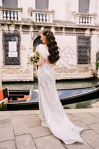 船站意大利威尼斯婚礼 一位穿着白色礼服的新娘 乘坐火车 手捧一束白色和红色的玫瑰 站在狭窄的威尼斯运河中停泊的贡多拉附近的码头上桉树背景