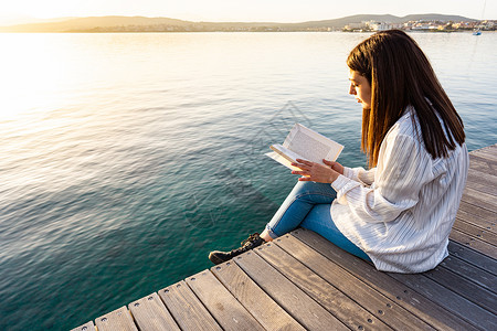 打码透明素材年轻女子在日落或日出时坐在码头上 在透明的海水上看书 女学生在大自然中学习大学课程 喜欢浪漫故事的书迷背景