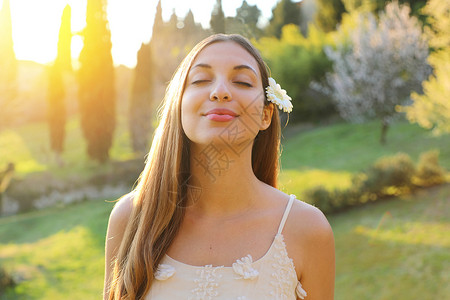 放平心态年轻微笑的女人闭着眼睛 耳朵上放着花深呼吸的画像 积极的春姑娘情感面孔表达感受生活感知成功和平心态概念 自由快乐的女孩享受大自然背景