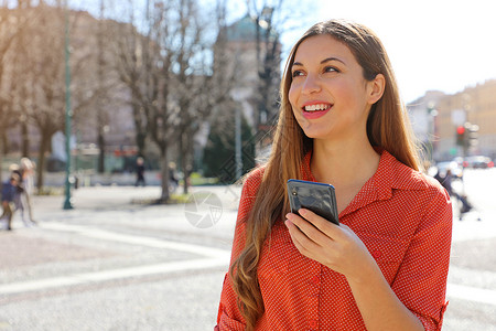 府前街一位自信的巴西女性手持手机在市街一边看一看前视线肖像 照片来自Frontview城市白色电话微笑女士幸福牙齿女孩细胞街道背景