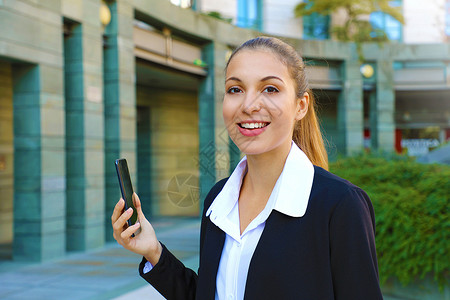 快乐的年轻成功女商务人士在外面使用手机 美丽的年轻女子穿着办公室西装 拿着智能手机 对着相机微笑 移动互联网概念背景图片