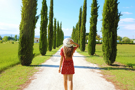 红色的柏树在托斯卡纳度假 穿着红色连衣裙和帽子的年轻快乐女人走在拥有典型托斯卡纳风景的小路上 女孩欣赏柏树胡同的景色背景