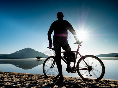 骑脚踏板在海滩上骑自行车的车手 后视人 用碎石脚踏板站立着护目镜爬坡车轮乐趣轮子运动风景日落眼镜太阳背景