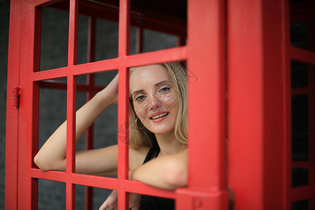 身穿黑色连衣裙的美丽金发女孩的画像站在红色电话亭里 靠在黑墙上 作为户外肖像时尚姿势电话微笑王国游客手机盒子英语旅游街道女士背景图片