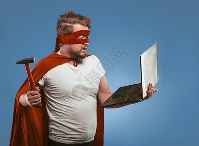 黑客服装素材超级英雄男子将用锤子砸碎或修理笔记本电脑 Denim Blue背景的红色超级英雄制服蒙面人侧视镜 计算机黑客概念背景