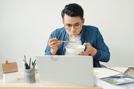 年轻的亚洲建筑师在办公室吃面面 在台式桌上职业桌子项目技术电脑面条男性成人自由职业者创造力背景图片