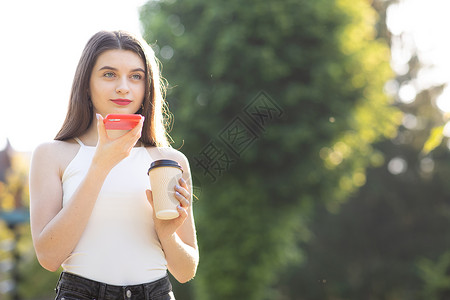 AI文字素材在公园散步的女孩用智能手机说话 快乐的女孩使用智能手机语音识别音频 ai 消息语音功能在线行走在公园 她手里拿着一杯咖啡背景