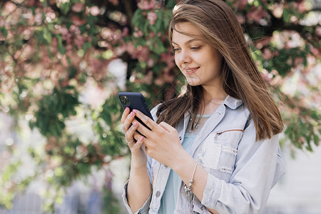 短信提示在户外用手机打字的快乐时髦女人的画像 开朗的女孩走在城市背景下的智能手机 微笑的女士在外面手里拿着手机肖像动作性格电话短信女性街背景