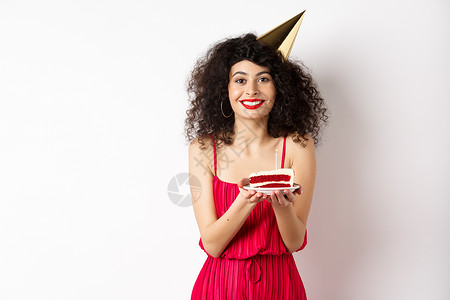 生日连衣裙穿着红色连衣裙 戴着派对帽庆祝生日 拿着生日蛋糕许愿 对着镜头微笑 站在白色背景上的漂亮女人女士黑发浪漫促销嘴唇投标裙子魅力工作背景