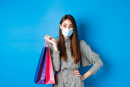 Covid19 大流行病和生活方式概念 令人惊讶的妇女在购物时佩戴医疗面具 拿着袋子和看相机 蓝背景工作室幸福社交女性广告卫生蓝背景图片