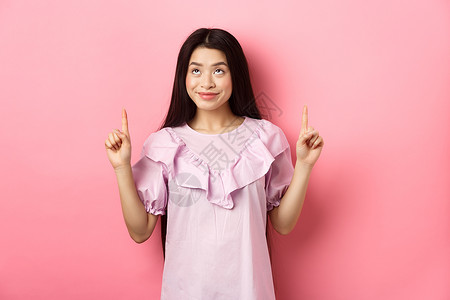 美丽的笑笑的亚洲少女女孩 举起手指 看着广告 面脸快乐 站在粉红背景上站着女性购物女士化妆品学生投标工作室青少年情感促销背景图片