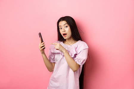 粉色年轻情感看书兴奋的美丽亚洲女性 指着智能手机 看起来很好奇 查看在线促销 站在粉红色背景下互联网上网女孩购物阅读投标电话女士青少年化妆品背景