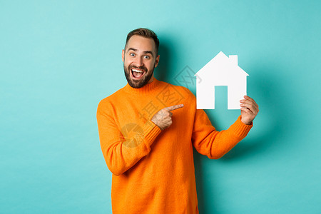 地产概念 快乐的男子用手指对着纸牌房子和微笑 站立在浅蓝背景上工作室毛衣胡子标识潮人促销横幅情感广告房地产背景图片
