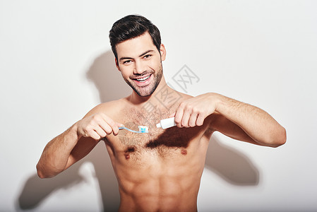 年轻的黑头发小黑头发男人 站着不穿衬衫 挤牙膏 在白背景上孤立的牙刷上背景图片