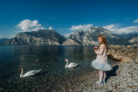 天鹅公主一个穿着漂亮的白色连衣裙的小女孩沿着加尔达湖的堤岸散步 一个女孩在意大利的一座山和湖泊的背景下被拍照 Torbole微笑礼服公主背景