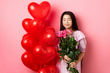 心和气球情人节的概念 浪漫的亚洲少女梦想着爱情或约会 闭上眼睛微笑 手捧情人的鲜花 收到一束玫瑰和红心气球 粉红色背景花束女朋友女孩青少背景