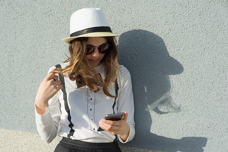 戴帽子的少女户外肖像 女孩使用智能手机 读写信息 复制空间 灰色文字墙背景背景图片