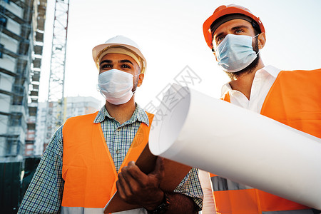 两名身穿工作服和医疗面具的男子与标物蓝图一起工作商业安全帽讨论伙伴制造业项目团队建筑学工程安全背景图片