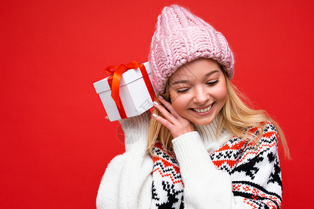 身着冬季毛衣和粉红色帽子的迷人正面微笑的年轻金发女性被隔离在红色背景墙上 她拿着带红丝带的白色礼盒 俯视着背景图片