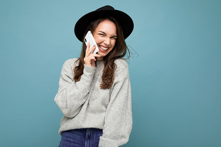 戴着黑帽和灰色毛衣的迷人年轻笑笑女人手持智能手机 看着背景中突显的相机背景图片