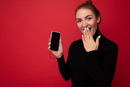 三品一标照片中 身穿黑色毛衣 身穿黑色毛衣的黑发年轻女性惊讶地站在红色背景中 她的照片上显示的手机屏幕空空如也 可以模拟看着相机背景