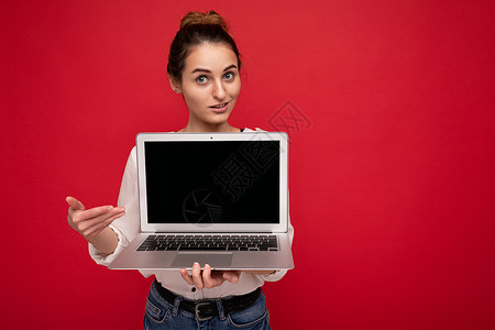 紧贴着美丽 微笑的年轻快乐女子的近距离肖像 她拿着电脑笔记本电脑看着照相机 用红色墙背景隔绝的穿便衣智能服装露脸背景图片