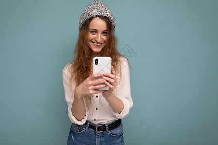 蓝色背景墙上隔着蓝色背景墙隔开的迷人 积极的年轻金发卷发女性照片 她拿着电话 使用在线交流看着相机背景图片