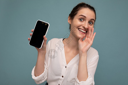 三品一标美丽积极的年轻金发卷发女性穿着休闲白衬衫 与世隔绝在蓝色背景墙上 拿着手机 展示带有空屏幕的智能手机 用于模拟看着相机和说秘密幸背景