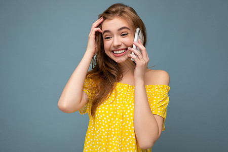 三品一标身穿时尚黄色夏装 面带微笑 迷人的金发女郎站在蓝色背景中 与世隔绝 手持手机 边看边说话工作室短信幸福屏幕技术工具约会联系电话横背景