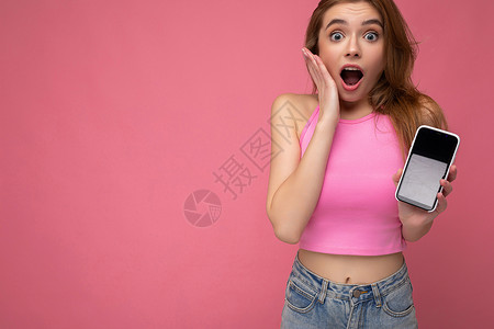 哇哦特写照片 令人惊奇的美丽快乐的年轻金发女子穿着粉红色的上衣 在粉红色的背景中与世隔绝 手里拿着空的空间 并展示带有空显示屏的手机背景